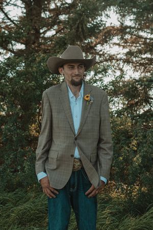Groom in plaid blazer, cowboy hat, jeans, and sunflower boutonniere standing outdoors among green trees.