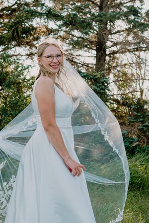 Smiling bride wearing glasses in white dress with lace veil in outdoor setting.