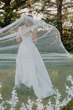 Bride in white gown with flowing lace veil billowing in the wind outdoors.