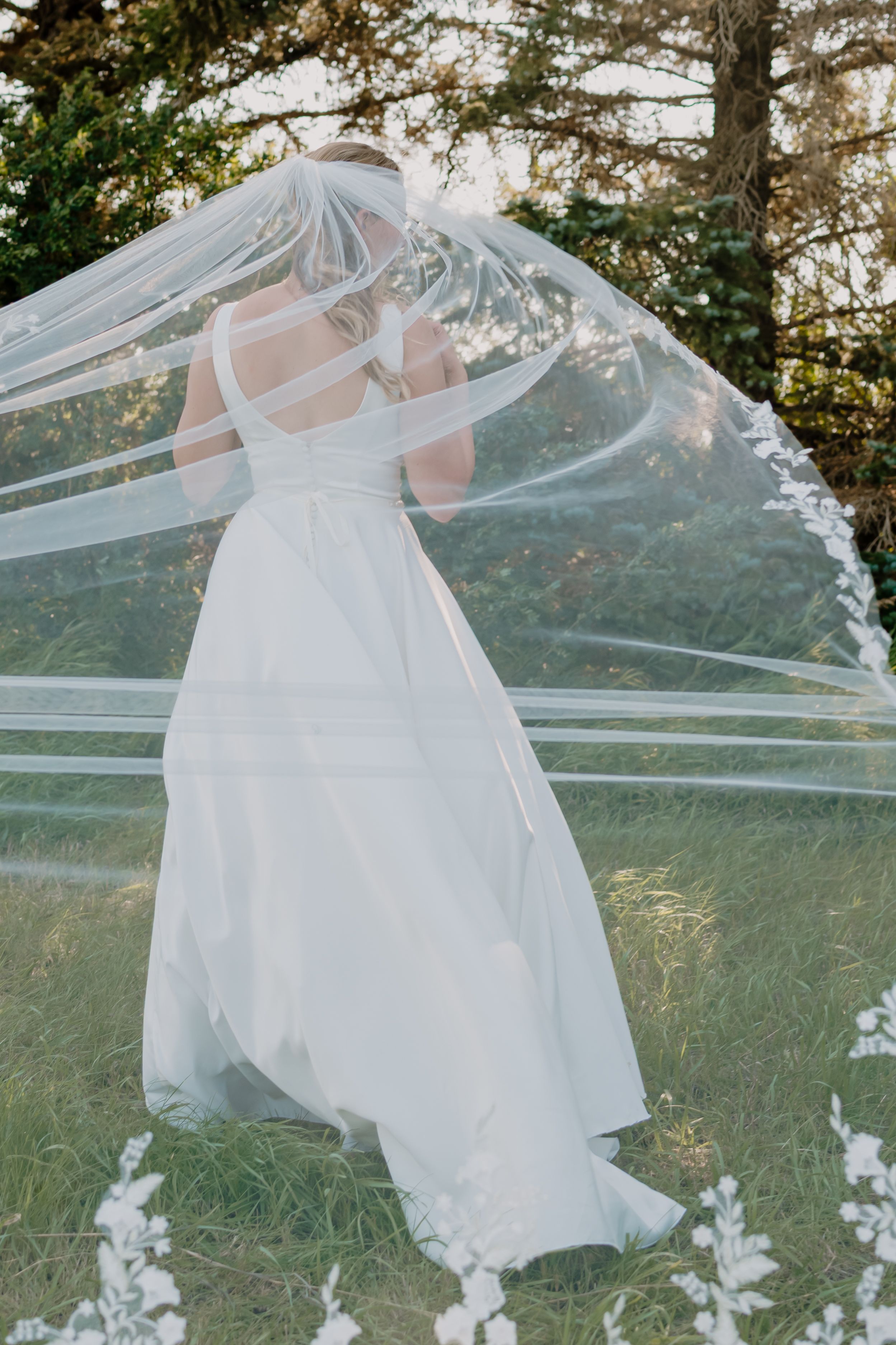 Bride in elegant white wedding dress with flowing veil billowing in the breeze outdoors.