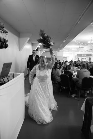 Bride in lace wedding gown raises bouquet while entering reception hall in black and white wedding photo.