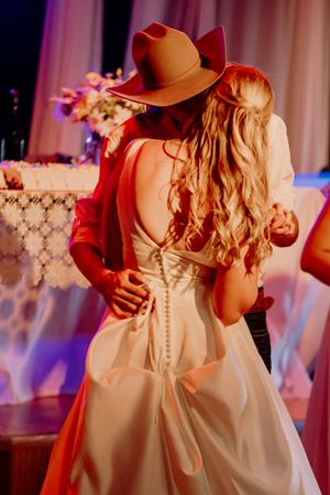 Bride in backless wedding gown and cowboy hat shares first dance with groom at rustic wedding reception.