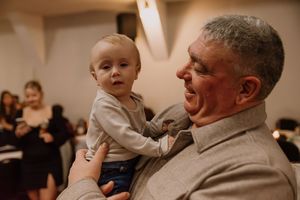 An elderly man joyfully holds a smiling baby at an indoor event, sharing a warm and tender moment.