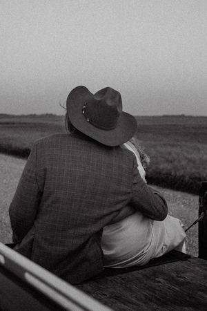 Couple embraces on rustic bench overlooking field at dusk, groom wearing plaid jacket and cowboy hat.