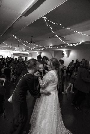 Black and white photo of bride and groom sharing a kiss with baby during wedding reception dance.