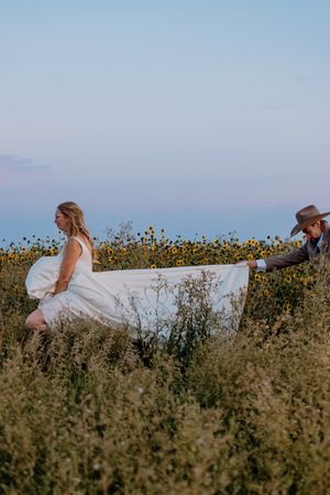 Bride in white gown walks through sunflower field while groom in cowboy hat carries her train at sunset.