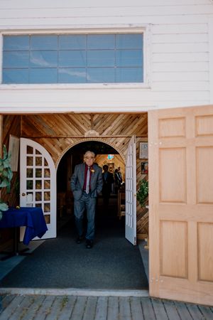 Man in suit walking through wooden arch doorway of a rustic wedding venue with white exterior walls.