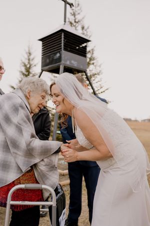 Bride in lace gown and veil shares a joyful moment with elderly grandmother at outdoor wedding ceremony.
