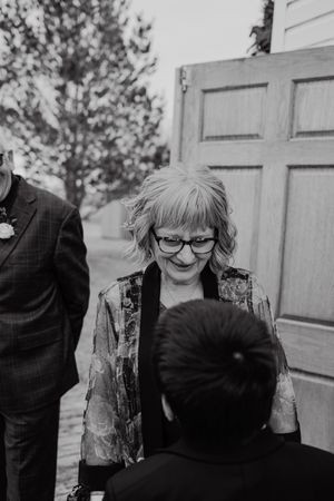 Black and white photo of a smiling woman with glasses and short blonde hair greeting a guest at an outdoor wedding event.