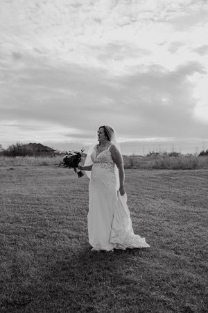 Black and white photo of a bride in a lace wedding gown holding a bouquet, standing in a field under a cloudy sky.