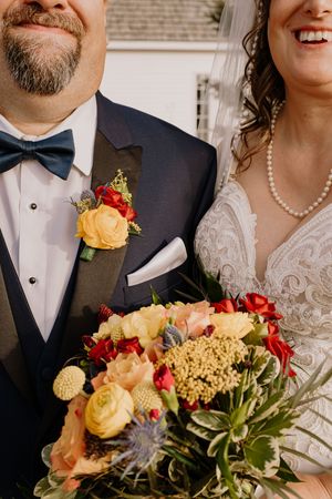 Bride and groom in wedding attire, groom in navy tuxedo with yellow boutonniere, bride holding colorful floral bouquet.