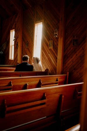 Bride and groom seated together at the altar of a rustic wooden chapel bathed in natural light.