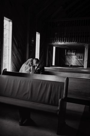 Black and white photo of a bride and groom sharing a kiss in a wooden chapel pew, illuminated by soft window light.