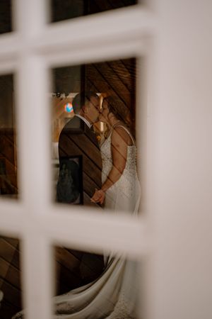 A bride and groom share a kiss, viewed through a white lattice window frame, with the bride's veil flowing gracefully.
