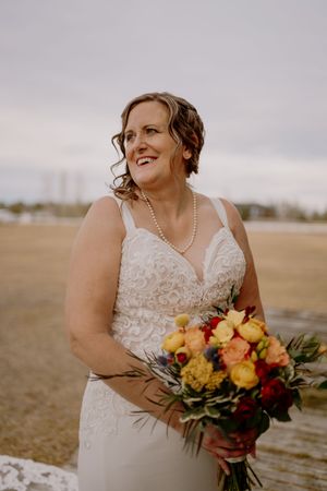 Bride in lace wedding dress holding colorful bouquet of flowers outdoors on overcast day.