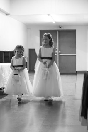 Two young flower girls in white dresses walk together in black and white wedding photo.