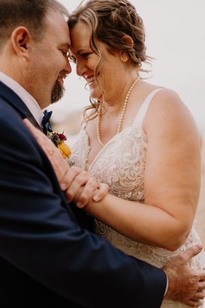 A bride in lace gown and pearl necklace shares an intimate moment with her groom in a navy suit during their wedding.