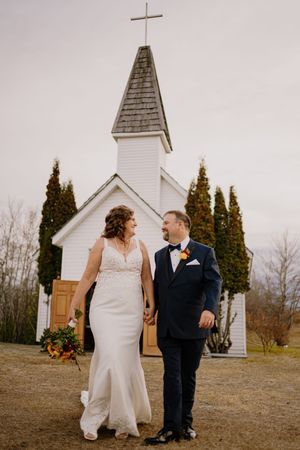 A bride and groom hold hands and smile at each other outside a white chapel with a cross on the steeple.