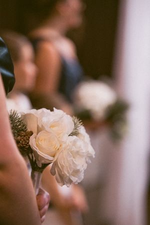 Bridesmaid holding a white peony and rose bouquet with pine accents during a wedding ceremony.