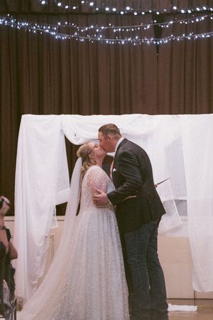 Bride and groom share their first kiss at the altar, decorated with white draping and twinkling fairy lights above.