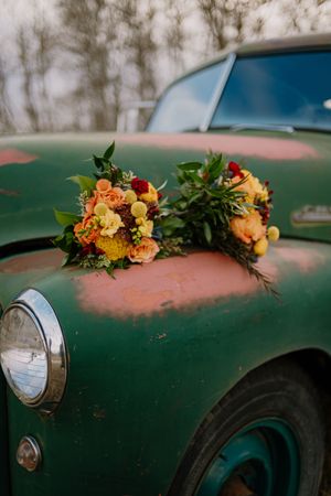 Two colorful wedding bouquets with orange, yellow, and red flowers resting on a vintage green rusty car hood.