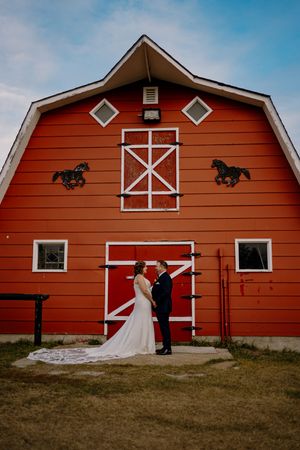 Bride and groom stand before a classic red barn with white trim and horse silhouettes on their rustic wedding day.