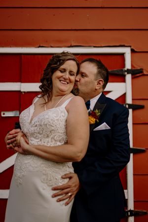 Bride in white lace gown smiles as groom in navy suit kisses her cheek before a red barn door.