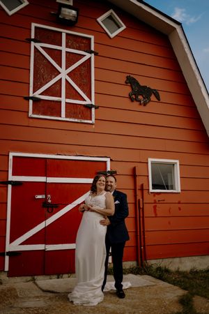 Bride and groom embracing in front of a red barn with white trim and horse decor at a rustic wedding venue.