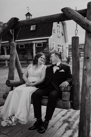 Black and white photo of bride and groom sitting on rustic log swing in front of a barn on their wedding day.