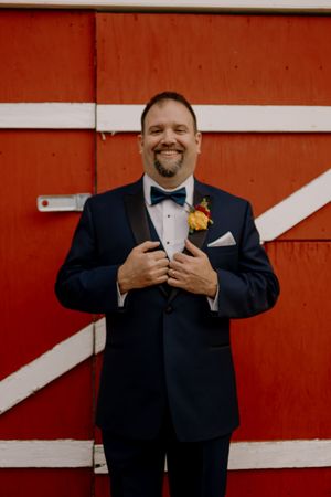 Smiling groom in navy tuxedo with bow tie and boutonniere poses in front of a red barn door.