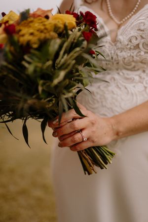 Bride in lace wedding dress holding a rustic bouquet of yellow and red flowers with greenery, diamond ring visible.