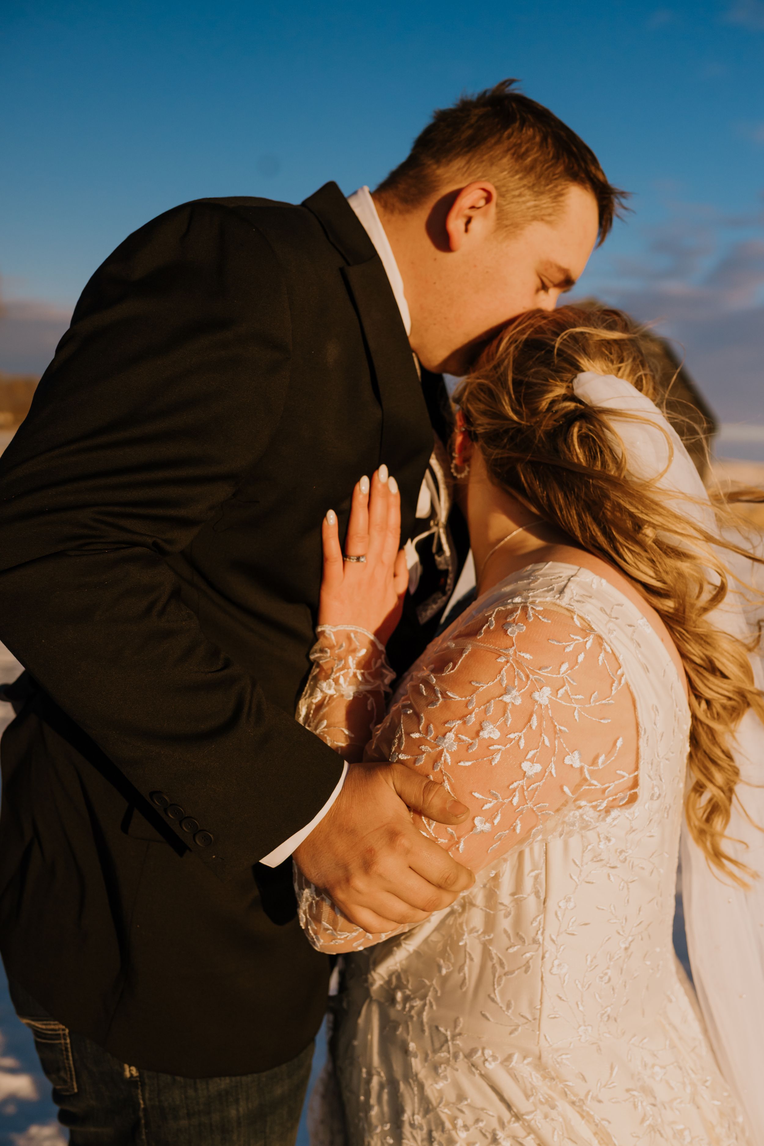Bride and groom share a romantic kiss outdoors at golden hour, with the bride in a lace long-sleeve wedding gown.