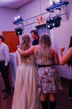 Two women in formal dresses dance together at a wedding reception under stage lights near a fire exit sign.