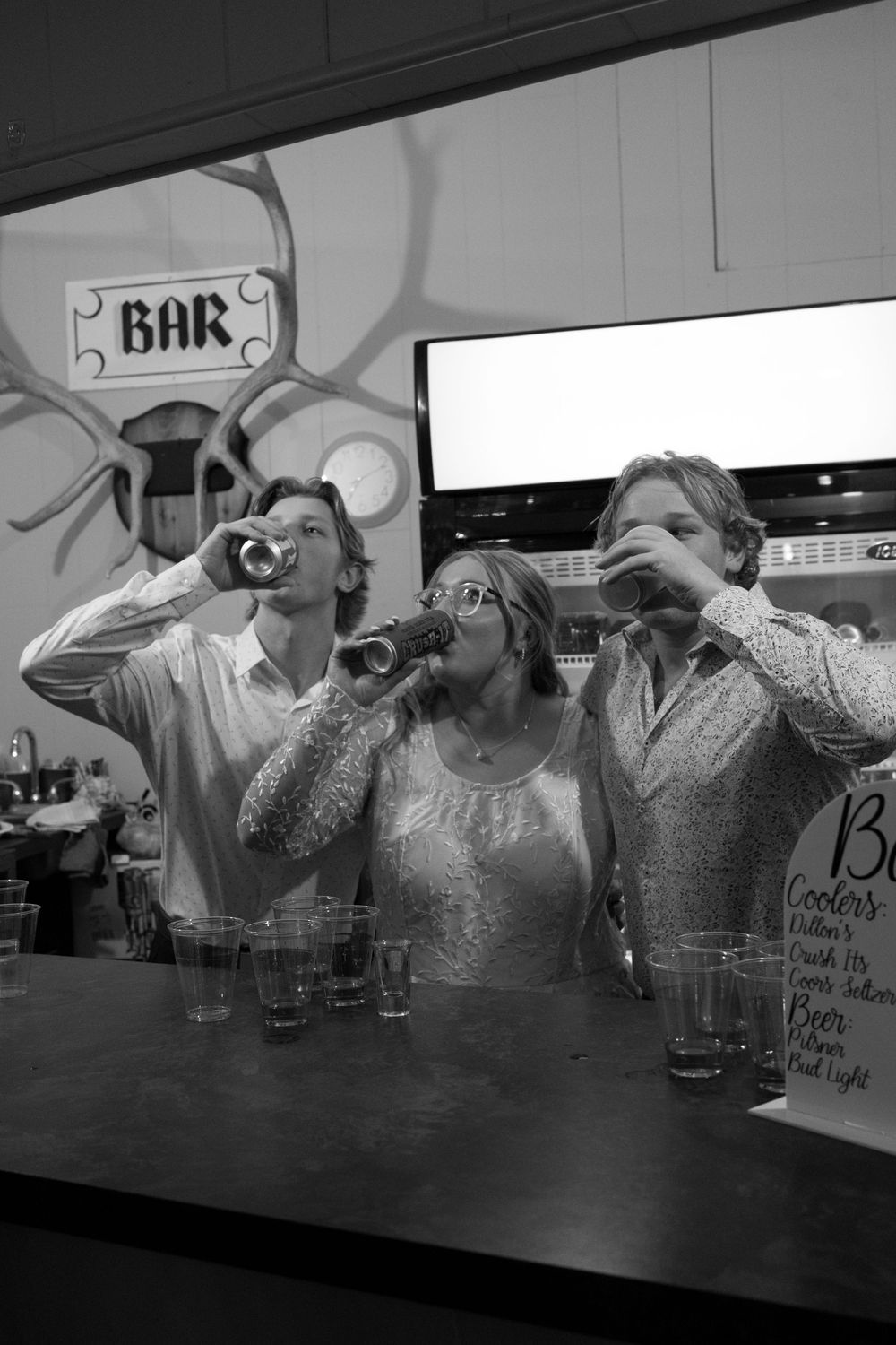 Three people drinking at a bar in black and white photo, two men and a woman raising glasses and bottles.