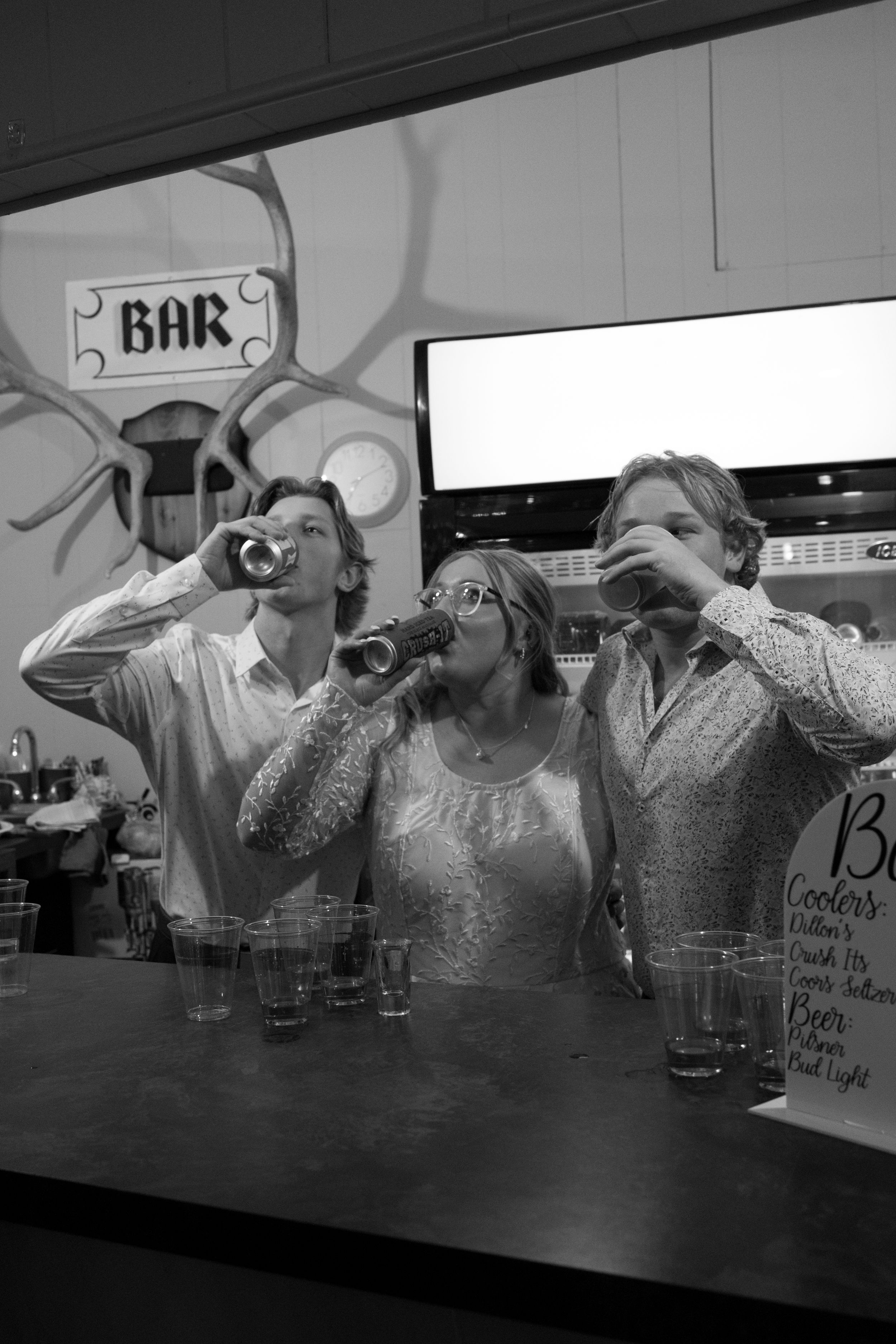 Three people drinking at a bar in black and white photo, two men and a woman raising glasses and bottles.