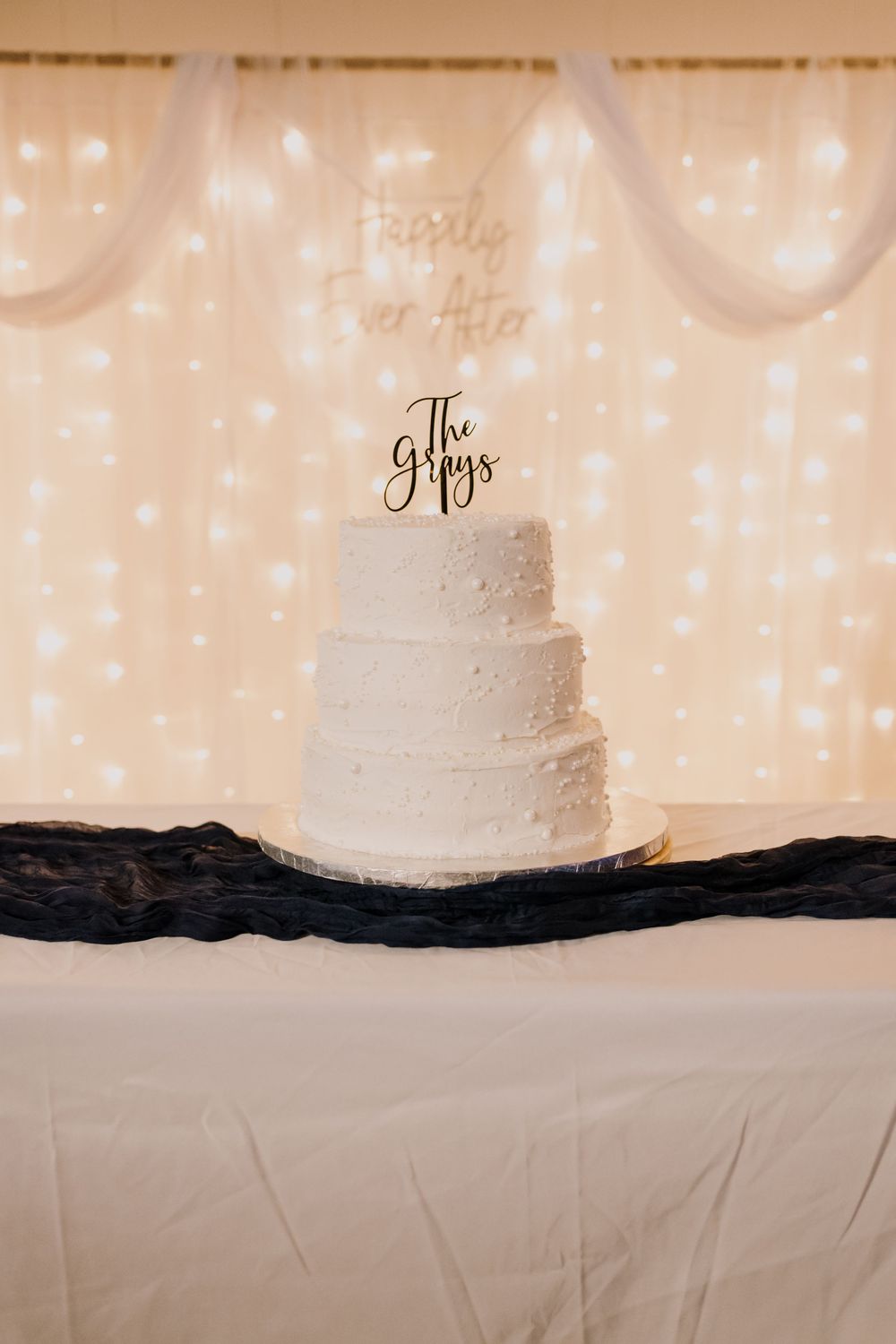 Three-tier white wedding cake with The Grays topper on a draped table, fairy light backdrop with Happily Ever After sign.
