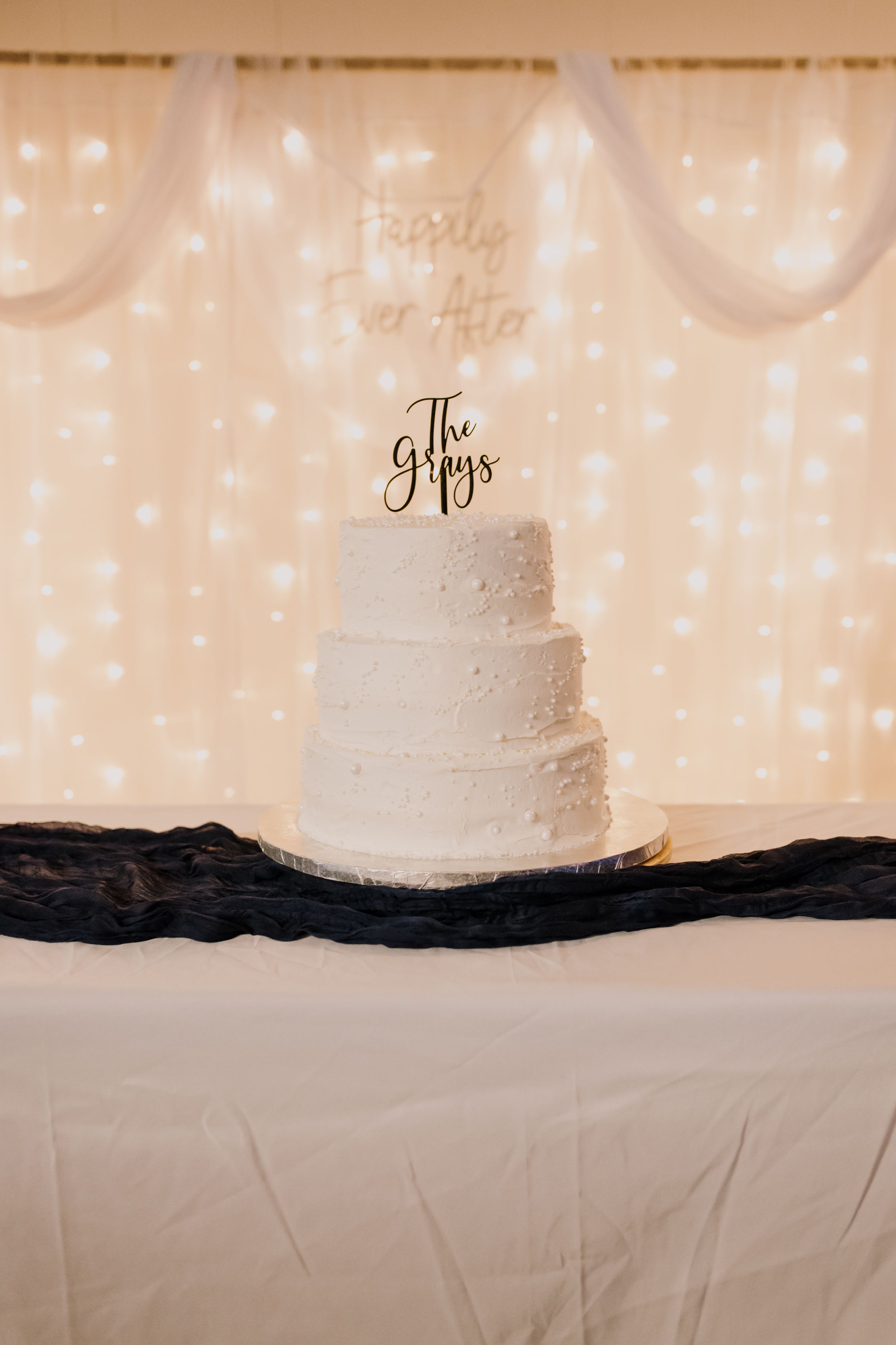 Three-tier white wedding cake with The Grays topper on a draped table, fairy light backdrop with Happily Ever After sign.