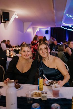 Two women smiling at wedding reception table with drinks and desserts under purple lighting.