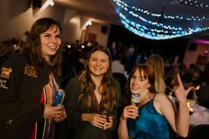 Three smiling women holding drinks at an indoor party with festive blue string lights in the background.