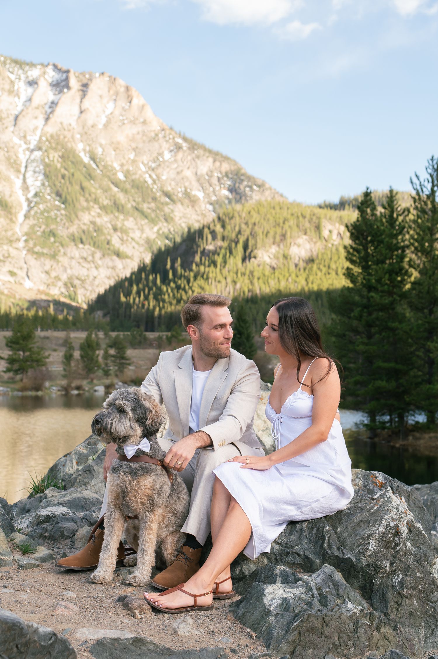 Couple sitting on rocks with fluffy dog, mountain lake and pine trees in background for an elopement at Officer's Gulch in Colorado.