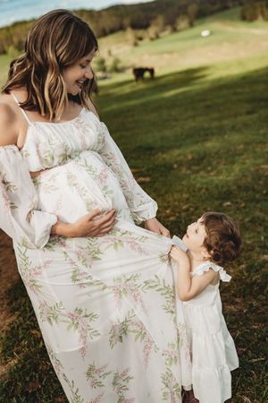 Pregnant woman in floral dress smiling at toddler girl outdoors on green hillside.