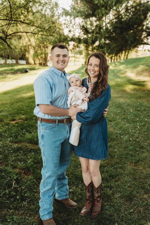 A smiling couple stands outdoors on green grass holding their baby, wearing casual denim outfits in a warm, sunny park setting.