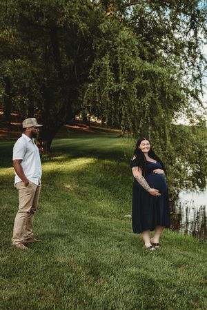 Pregnant woman in navy dress holds belly while man in white shirt watches, standing on green grass near a pond.