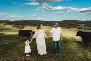 Pregnant woman in white dress with husband and toddler on green farm pasture with black cows and rolling hills.
