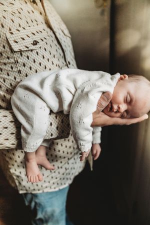 A newborn baby sleeping peacefully in white knit outfit, cradled in one hand by a parent wearing a patterned shirt.