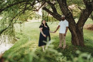 Pregnant couple holding hands under a large tree in a lush green park, woman in black dress, man in white shirt.