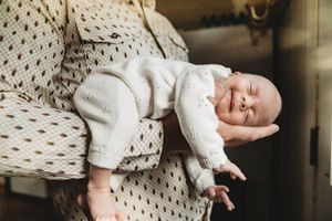 Newborn baby in white knit outfit sleeping peacefully in a caregiver's arms, soft natural light.