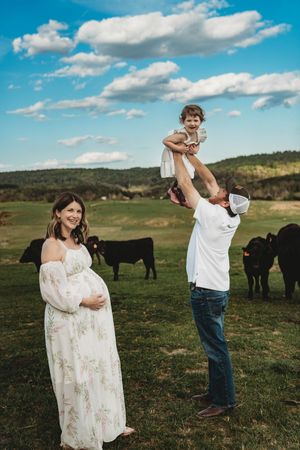 Pregnant woman in floral dress stands smiling as man lifts toddler in air on green farm with black cattle.