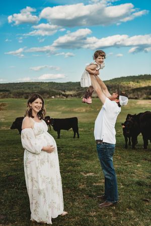 Pregnant woman in floral dress smiles as man lifts toddler girl in air on green farm with black cattle.