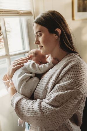 A young mother tenderly holds her sleeping newborn baby close to her chest near a bright window.